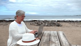 Smiling senior woman typing on laptop sitting at the beach working, shopping, writing blogs. Elderly lady enjoying outdoors at sea using modern technology. Horizon over sea - Powered by Shutterstock - Get 15% off with code: PIKWIZARD15