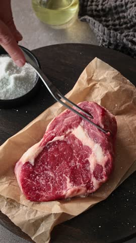Chef sprinkles salt on fresh raw beef steak in kitchen, close-up