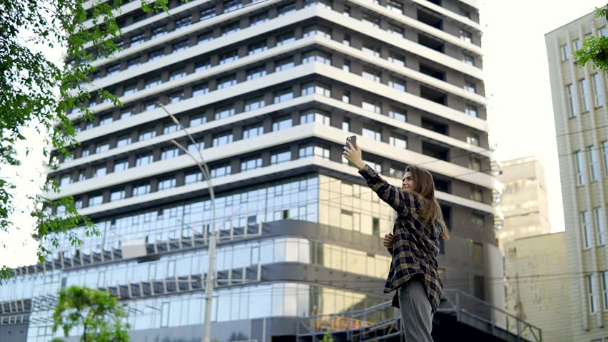 A young smiling girl presents a new housing complex over the phone. Rejoices and dances in front of the new house. Talking on the phone with a friend. Wearing a white T-shirt and a plaid shirt.