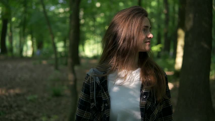 A young smiling girl with a beautiful smile was blinded by the sun in a green forest. She is wearing a white T-shirt and a plaid shirt. Athletic build.