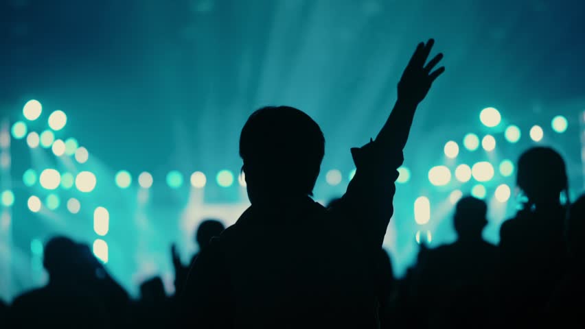 "A young woman is clapping her hands above her head at a Christian music festival. Many members of the audience are raising their hands and singing songs of praise to God with hope."