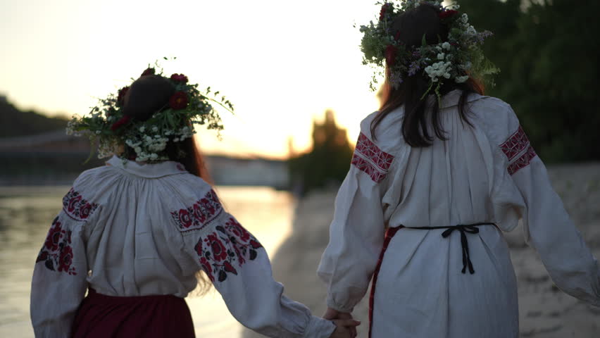 Slow motion. Rear view. Two women walk along the sandy beach of the Dnipro River during the celebration of Ivan Kupalo. The women wear traditional embroidered shirts and wreaths of wild herbs and