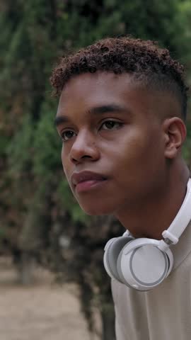 Young adult african american man with headphones sitting on bench outdoor. Millennial student boy waiting for someone in public park. Vertical HD video.