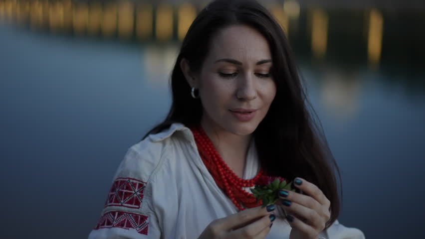 A woman in a traditional Slavic costume holds a red flower in her hands and puts it behind her ear, smiling, while standing outdoors on a summer evening