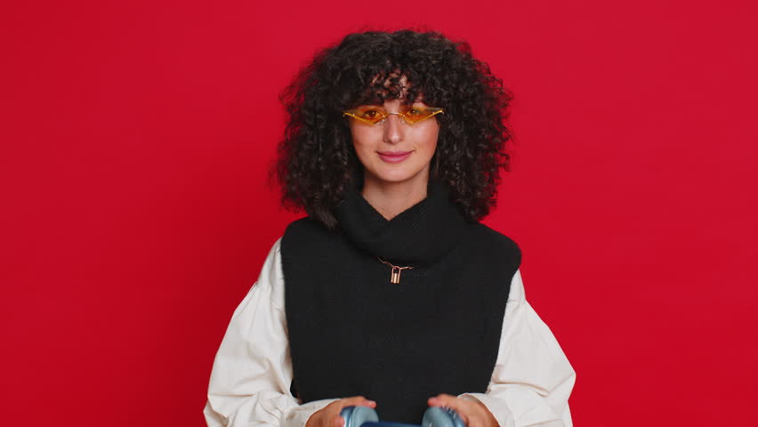 Caucasian young woman with curly hair listening music on headphones, dancing disco fooling around having fun expressive gesticulating hands. Music app advertisement. Girl isolated on red background
