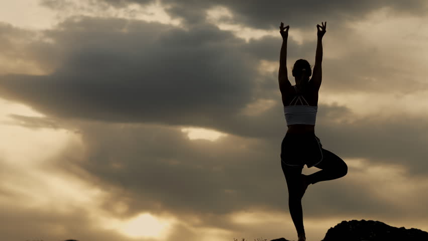 Silhouette of a woman practicing yoga tree pose on a mountain at sunset outdoors, expressing balance, mindfulness and peaceful nature atmosphere.