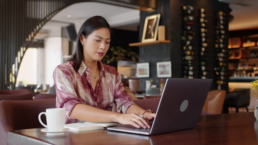 Medium shot of young Asian female entrepreneur making notes when working on wireless laptop in cafe