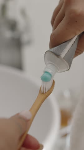 Vertical shot of hands of woman squeezing out toothpaste on bamboo toothbrush in bathroom