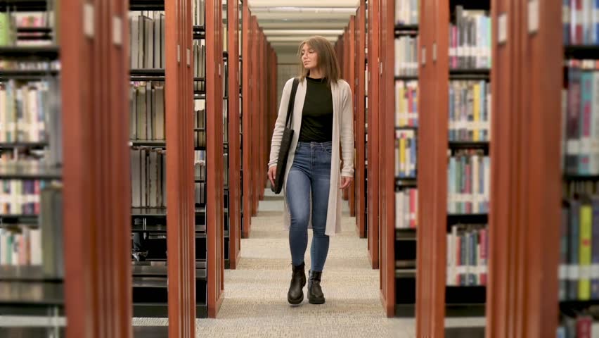 A young woman in casual attire walking through a library aisle filled with bookshelves, creating a serene and focused atmosphere