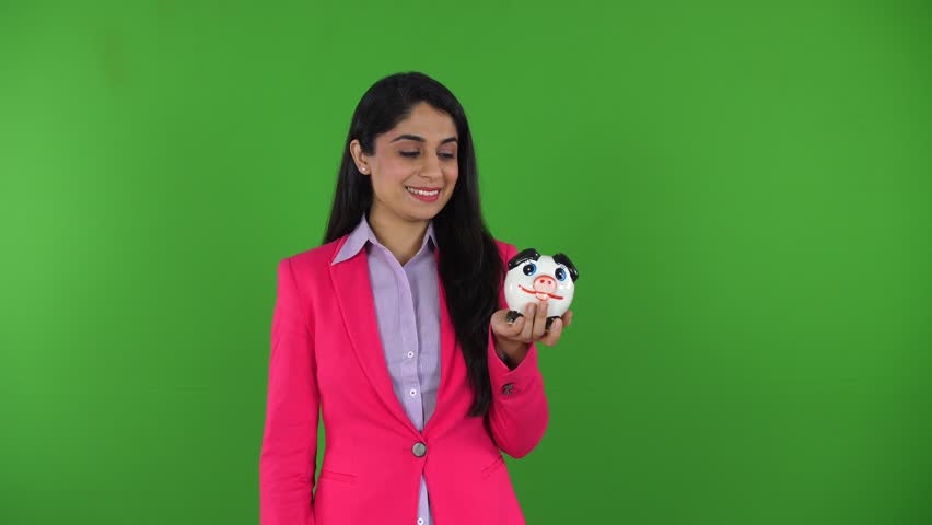 A smart Indian woman holding a piggy bank and pointing towards it against green screen - money concept, financial stability. A young woman with a professional look plans for future savings 