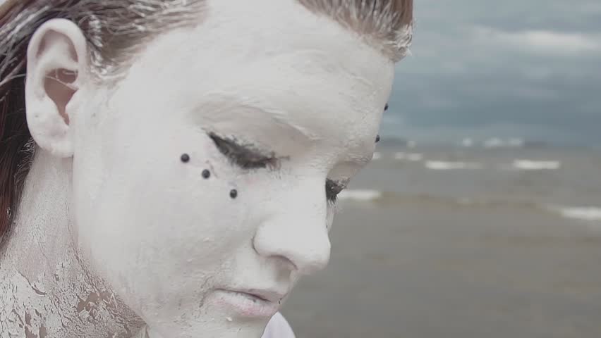 Portrait of young performance caucasian red haired girl artist covered in white paint shaking head on wavy sea shore, cold cloudy day
