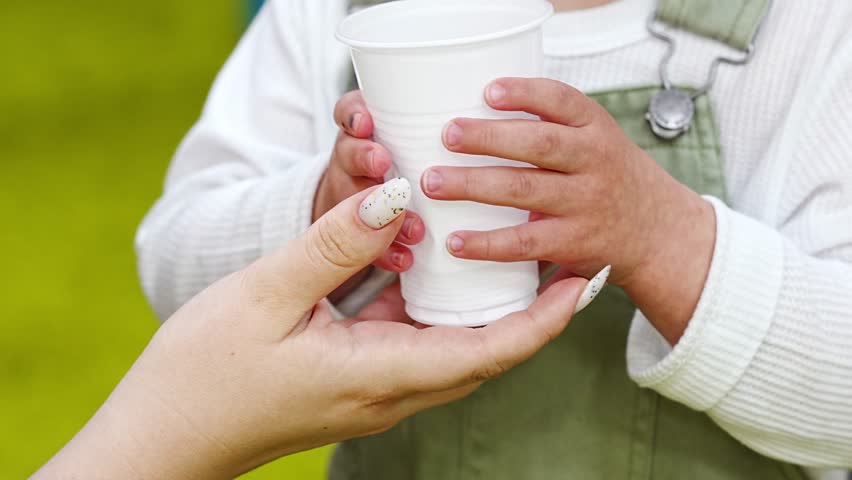 without a face. Mom helps a small child drink water from a plastic cup.