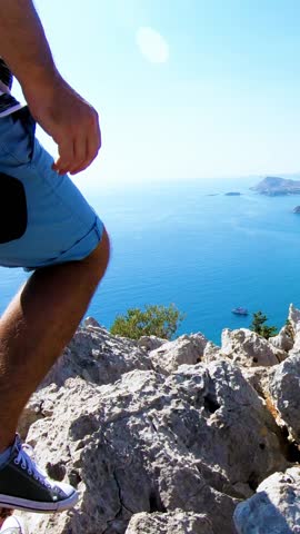 A young man reaching the top of the mountain and standing on cliff with arm raised. Mediterranean sea coast and rocks in the background