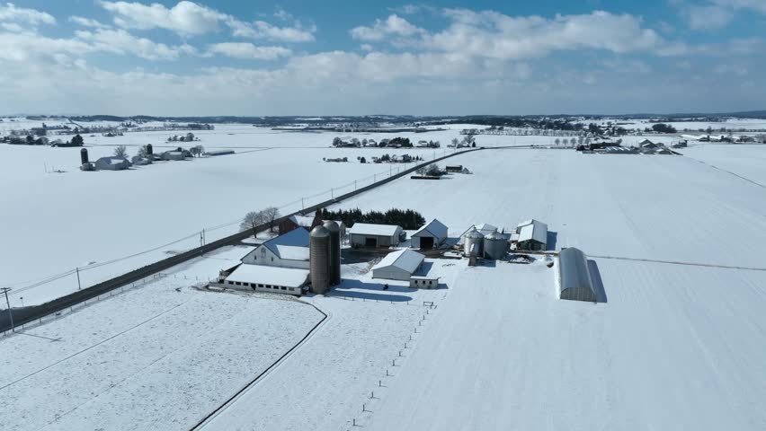 American farm in rural USA during winter. Farmland and fields blanketed in white snow. Sunny day. Aerial reveal.