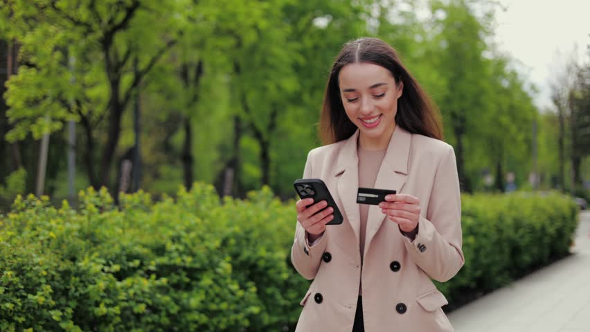 Happy young caucasian woman enjoying good weather with modern smartphone and plastic credit card in hands. Cheerful female engaging in online shopping process during walking time near public park.
