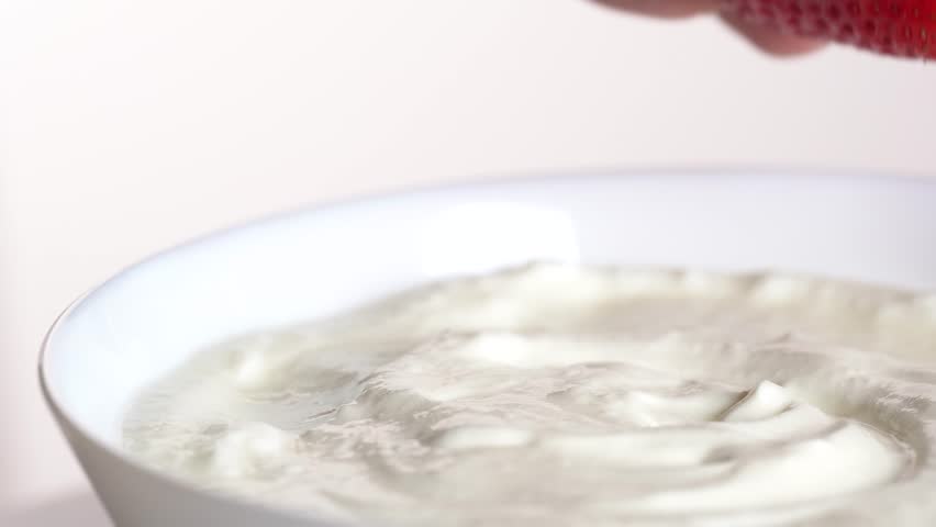 A hand immerses a ripe strawberry in white cream. Summer eating. Dipping fresh strawberries in Greek yogurt, macro.