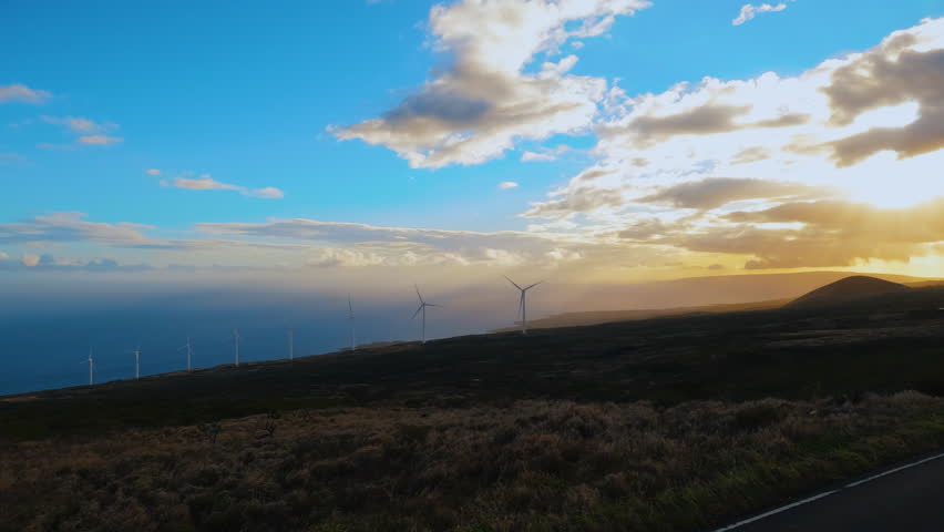 Driving near the wind farm at sunset. Pov footage of wind turbines at mountainous terrain near the ocean bay. Ride near Kaheawa Wind Power wind farm on Maui island, Hawaii. Road to Hana.