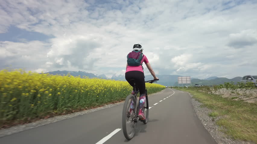 Female cyclist riding on a bike lane in the rural landscape on an overcast summer day, rear view. Cycling, nature, and recreation concepts.