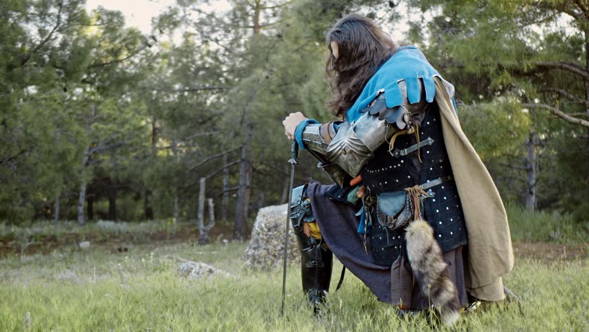 A man in a medieval knight costume stands kneeling and leaning on a sword stuck in the ground in front of him in the bright rays of the sunset. Close-up. Side view. Prayer. Historical reconstruction