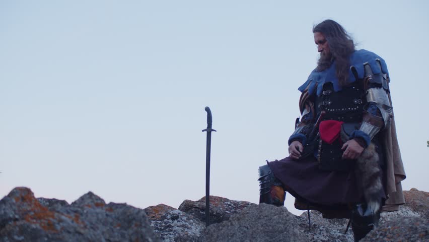 A man in a medieval knight costume pulls a sword from a stone against the background of a blue evening sky. Historical reconstruction. Costume performance. King Arthur