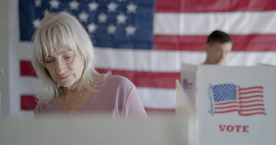 Elderly woman voter casts votes in election at booth, looks thoughtfully to one side, then puts on "I voted" sticker and smiles to camera. Asian voter, soft focus, behind her. US flag on back wall