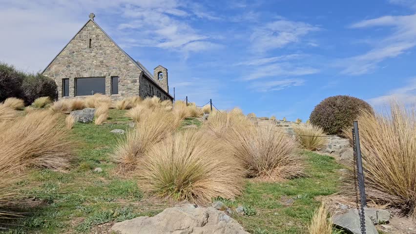 Young woman climbing up stairs towards a small anglican church, the church of Good Shepherd in the shores of Lake Takapō in South Island, New Zealand. Wind moving the long flexible twigs of bushes.