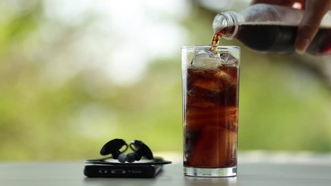 Hand pouring cola into glass with ice and Smartphones Headphones on the floor