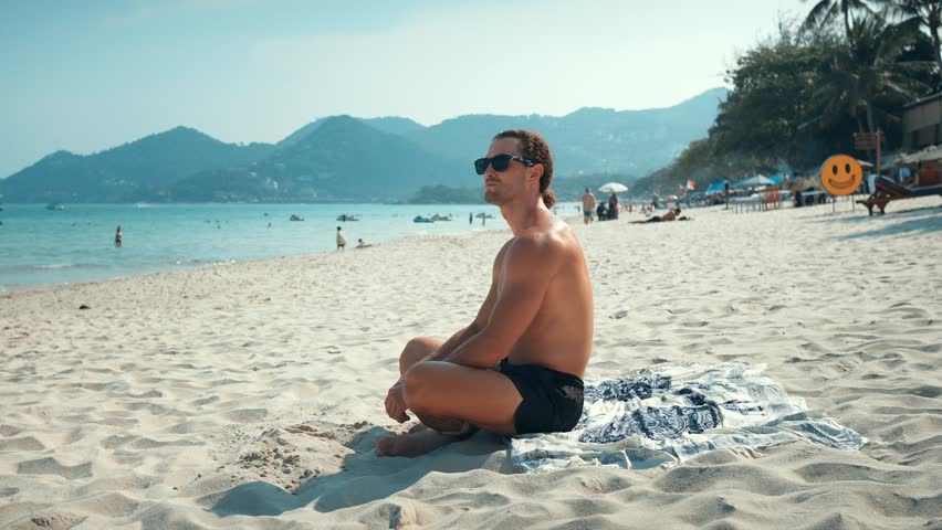 Millennial sitting sunbathing on beach and looking at seascape. Warm sand sound of surf and alight breeze from sea impress man with beauty. Calm man immersing himself process relaxation, enjoying sea.