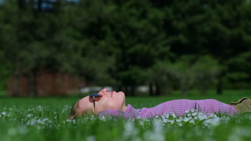 Close-up shot of relaxed woman laying on back on a flowering lawn in sunny day. Happy woman wearing sunglasses lies on a green grass in summer park.