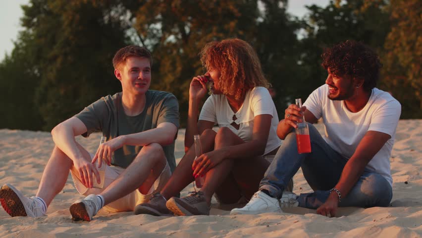 Three students telling funny situations in life when meeting the sunset. Best friends drinking alcohol on rivercoast. Summer time chill atmosphere. Humorous mates can't stop laughing