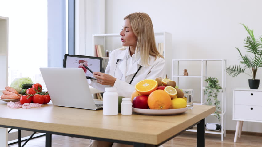 Caucasian doctor in white coat gives professional consultation remotely. Mature female nutritionist talks about the treatment of gastrointestinal tract to patient during online session.