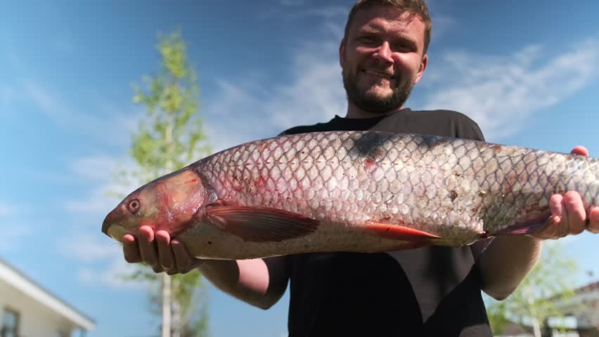 Satisfied young man with large freshly caught fish in hands stands outdoor demonstrating successful fishing. Low angle.  White Amur