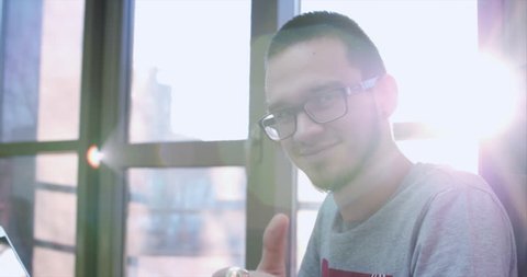 Portrait of a young happy man in glasses, businessman, programmer or trader, sitting in the office with laptop, looking at camera with sun shining behind, smiling happily, showing thumbs up. 4K