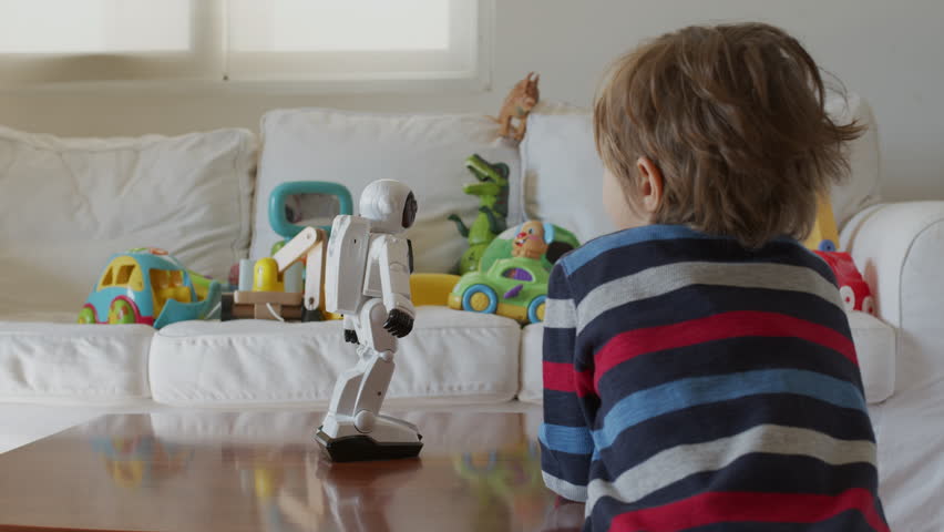 A young boy attentively observes a robot toy on a table, surrounded by colorful playthings in a well-lit living room.