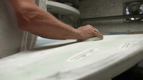 Man uses handheld palm sander to smooth the tail of a white surf board in workshop - close up shot - Powered by Shutterstock - Get 15% off with code: PIKWIZARD15