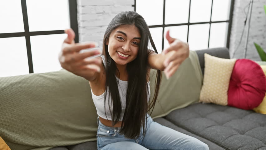 Cheerful young indian lady, with arms wide open for a warm hug, relaxing on sofa at home, welcoming happiness with a radiant smile, awaiting friendship love gesture indoors