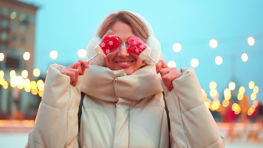 Joyful woman in earmuffs covering eyes with Christmas candies. Portrait of beautiful young female in warm clothes standing against blurred background with Christmas lights in city during evening.