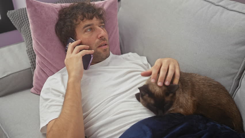 Stressed young hispanic man talking on the phone while petting his cat in a cozy living room.