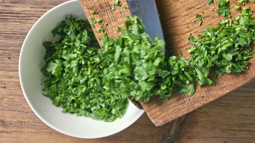 Chef puts fresh green chopped parsley leaves in bowl from wooden cutting board to prepare healthy food, top view. Food close up