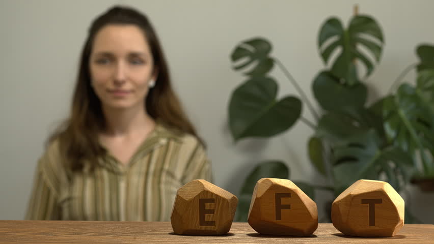 Letters EFT writen on irregular wooden blocks. Female tapping side of the eye (SE) meridian point in blurred background. Emotion-focused therapy treatment concept.