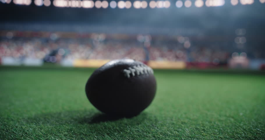 Close Up View of an American Football on a Green Field, Illuminated by Stadium Lights with Cheering Crowd of Fans in the Background. Atmosphere of a Night Time Football League Game