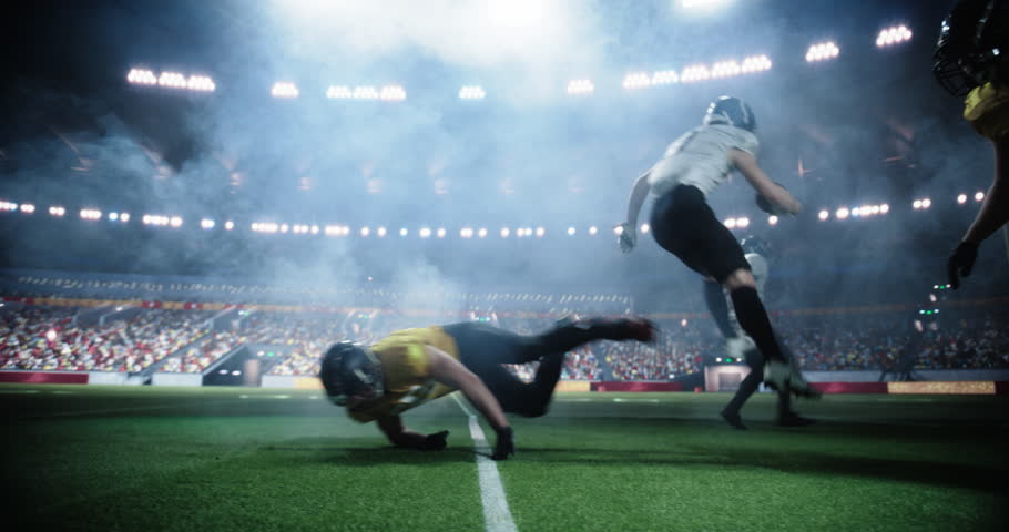 American Football Players Compete in a Stadium, Illuminated by Bright Lights With a Cheering Crowd in the Background. Athletes Play an Intensive Dynamic Game, Tacle, Try to Score a Touchdown