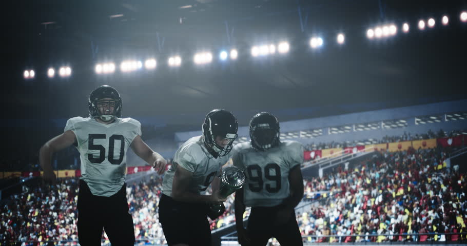 Joyful American Football Players Excited After Winning a Championship Match Trophy. Diverse Group of Athletes Standing Stadium Under Falling Confetti, Lifting the Cup in the Air
