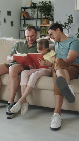 Vertical full shot of modern gay couple spending time with their daughter sitting on sofa in living room reading novel