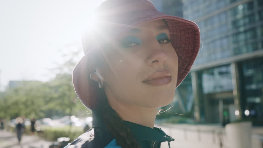 Portrait creative young woman hip-hop dancer posing looking at camera standing in city downtown, 80s 90s urban style retro outfit, blue jacket bomber and bucket hat, sunlight, sun beams bright flares