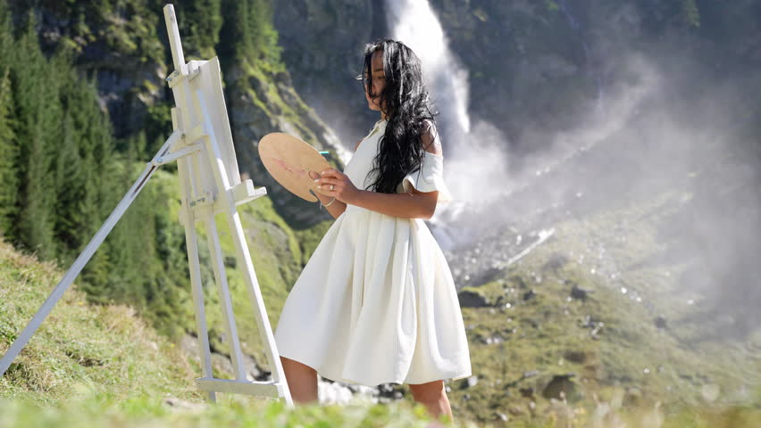 Female artist painting an alps landscape. Artist woman drawing outdoor. Beauty woman artist draw pictures in waterfall. Female artist paints a picture on canvas. Painting Outdoors.
