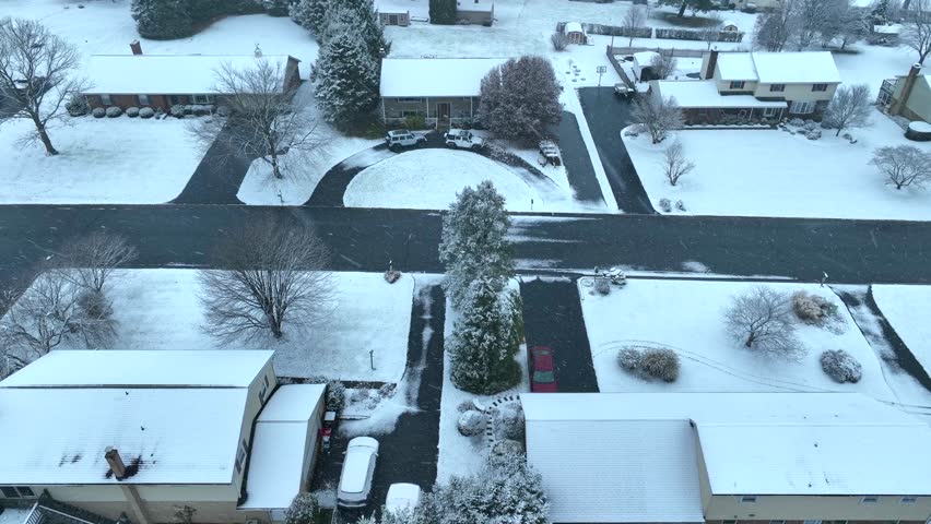 Winter view of a suburban neighborhood with snow-covered houses, yards, and roads, under a grey sky. Top down aerial truck shot during snow flurries.