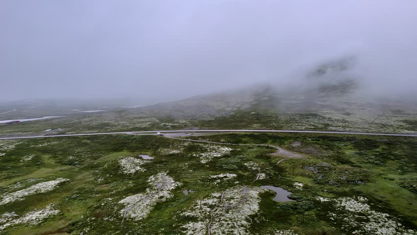 Flying over the green tundra and above a parking lot in Rondane National Park, near Venabygd, Norway, showcasing the misty nature landscape and disappearing into the cloudy mountains