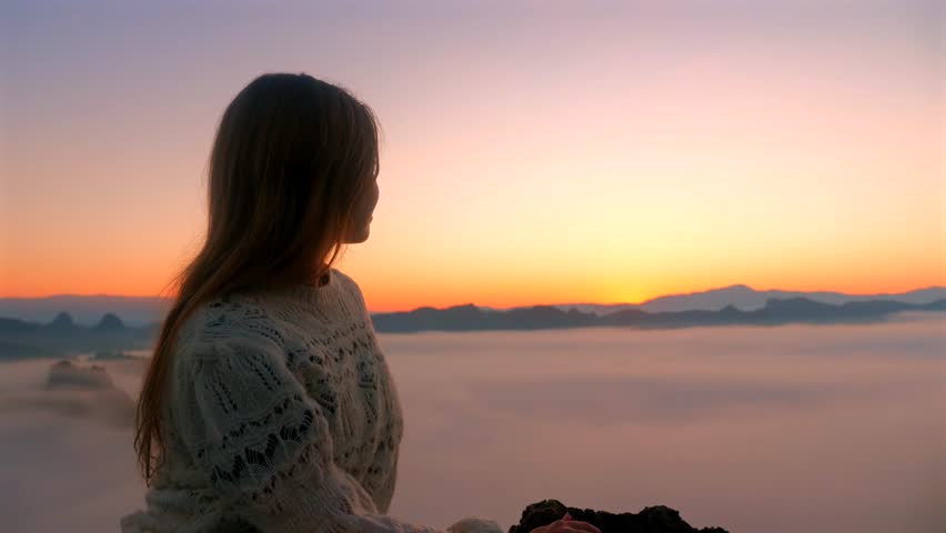 Woman enjoying panoramic sunrise view on mountain top with misty fog covering valley below and wearing cozy sweater in natural scenic outdoor landscape. Nature and travel exploration.