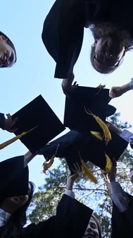 Classmates in graduation gowns toss their hats outdoors. Bottom view. Vertical video. 